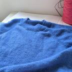 Blue knitted sweater on a white surface with a pink yarn ball in the background